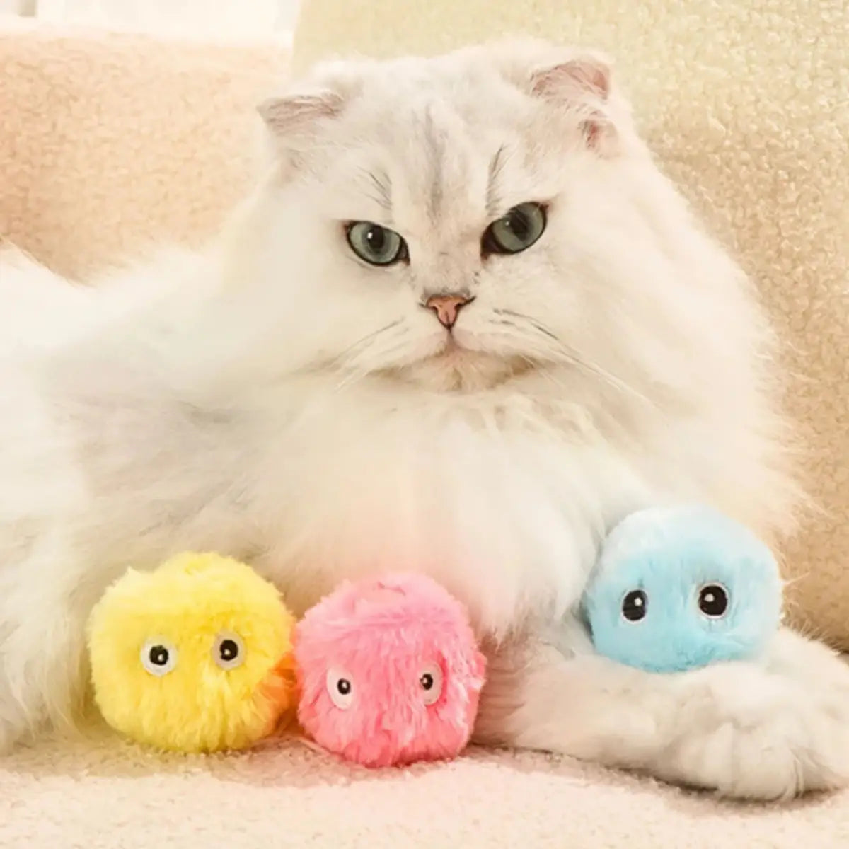 Chat blanc avec trois jouets peluches colorés Plush™ posés sur une couverture.
